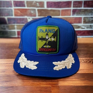 May include: A blue trucker hat with a mesh back. The front of the hat features a green, yellow, and black patch with a plane and the text "PAANG DRAGONFLY".