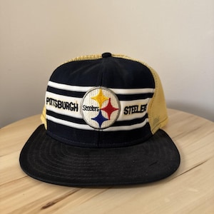 Puede incluir: Una gorra de béisbol de los Pittsburgh Steelers en negro y dorado. La gorra tiene una visera negra, una corona azul marino con rayas blancas y una parte trasera de malla amarilla. El logotipo del equipo está centrado en la parte delantera.