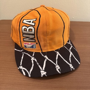 以下が含まれることがあります： オレンジと黒のNBAバスケットボールキャップ。キャップには、NBAのロゴが白と赤で表示され、クラウンにはバスケットボールのデザインが施されています。つばは黒で、白いネット模様です。