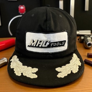 May include: Black baseball cap with the "MAC TOOLS" logo on a white patch. The cap has two embroidered leaf-like designs on the brim. The cap is made of a durable fabric.