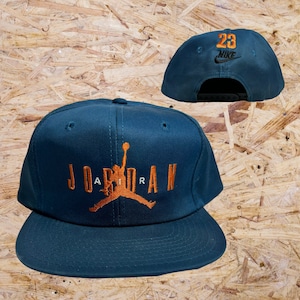 May include: A dark teal blue baseball cap with a black and white Nike logo and the number 23 embroidered on the front. The back of the cap has the word "JORDAN" embroidered in orange with a basketball player silhouette.