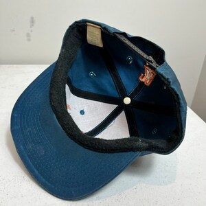May include: A blue baseball cap with a black brim and a white mesh lining. The cap has a brown embroidered logo on the front.