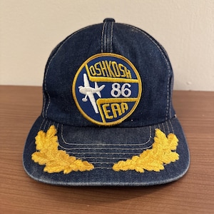 Può includere: Un cappello da baseball in denim blu con una toppa ricamata in oro che dice "Oshkosh 86 EAA" e un aereo bianco. Il cappello ha anche foglie ricamate in oro sul bordo.