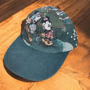 Puede incluir: Gorra de béisbol verde azulado con visera curva. La corona de la gorra presenta un diseño tejido con los personajes de Mickey y Minnie Mouse en una escena temática de golf. La gorra está hecha de tela texturizada.