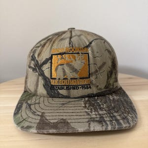 May include: Camouflage baseball cap with the Rocky Mountain Elk Foundation logo embroidered on the front. The logo features an elk silhouette in a square frame, with the text "ESTABLISHED 1984".
