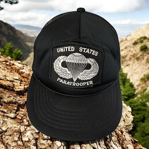 May include: Black mesh trucker hat with a black patch that says "UNITED STATES PARATROOPER" and features a silver embroidered parachute.