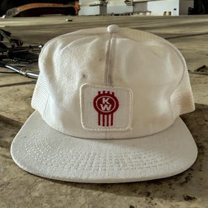 May include: White trucker hat with a red and white KW logo patch. The hat has a mesh back and a white brim.