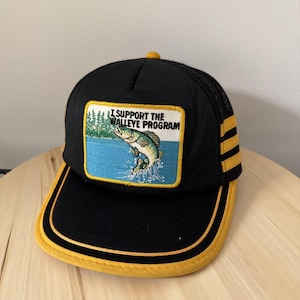Peut inclure: Casquette de camionneur noire avec une visière et des accents jaunes. L'écusson avant représente un poisson sautant au-dessus de l'eau bleue avec le texte "I SUPPORT THE WALLEYE PROGRAM". La casquette a un dos en filet et un bouton supérieur jaune.