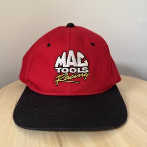 Może przedstawiać: Czerwono-czapka bejsbolowa z czarnym daszkiem i napisem "MAC TOOLS Racing" wyhaftowanym z przodu. Czapka ma zakrzywiony daszek i czarny guzik na górze. Czapka jest prawdopodobnie wykonana z wytrzymałej tkaniny.