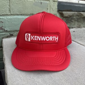 May include: A red trucker hat with a curved brim and mesh back. The front panel features a white rectangular patch with the word "KENWORTH" in white block letters and a logo.