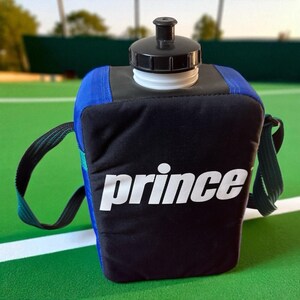 Puede incluir: Un portabotellas de agua negro y azul con tapa negra y botella blanca. La palabra "prince" está impresa en blanco en la parte delantera. El portabotellas tiene una correa verde y negra para llevarlo.