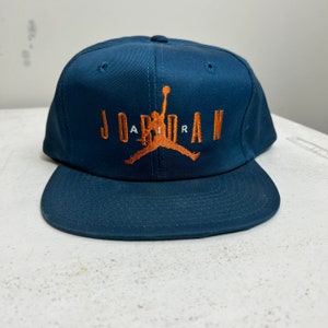 May include: A navy blue baseball cap with a white "AIR" and orange "JORDAN" logo featuring a basketball player silhouette.