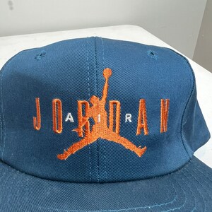 May include: A navy blue baseball cap with an orange embroidered Jordan logo and the word "JORDAN" in orange lettering.