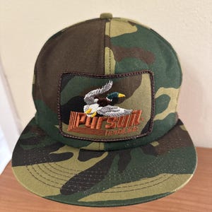 Könnte beinhalten: Camouflage-Baseballkappe mit einem braunen Patch, auf dem ein fliegender Enten und der Text "Pursuit Heritage" zu sehen sind.