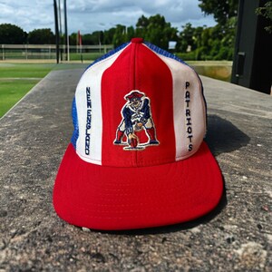 May include: A red, white, and blue baseball cap with a New England Patriots logo. The logo is a cartoon of a football player in a blue uniform. The cap is a trucker style with a mesh back.