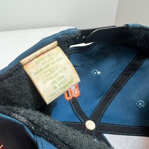 May include: A blue baseball cap with a black adjustable strap and a white button on the top. The cap has a black and white embroidered design on the front and a label on the side that reads "S20811WDI 560108 machine wash cold water 30 adult:one size fits all 100% Cotton RN #56323 made in TAIWAN RO.C".