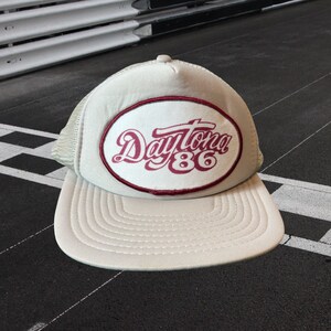 May include: A beige trucker hat with a red oval patch that says "Daytona 86" in white cursive lettering.