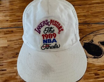 May include: White baseball cap with a patch that says "The 1989 NBA Finals Lakers Pistons".