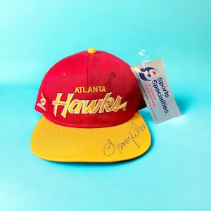 May include: A red and yellow baseball cap with the Atlanta Hawks logo and a signature on the brim. The cap has a tag attached that reads "Sports Specialties, The Leader in Sportswear".