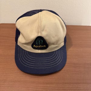 Puede incluir: Gorra de camionero beige y azul marino con el texto "Agratech" en un parche verde y negro. La gorra tiene una parte trasera de malla y una visera curva.