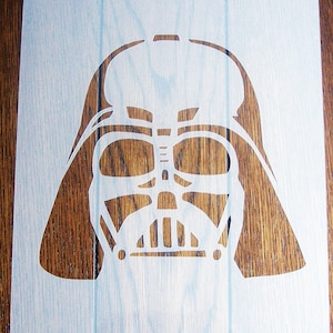 May include: A white stencil of Darth Vader's helmet on a wooden background.