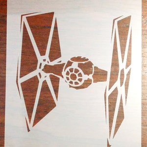 May include: White vinyl stencil of a TIE fighter from Star Wars. The stencil is on a brown wooden surface.