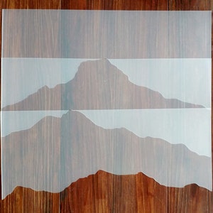 May include: Three clear plastic stencils of mountain ranges, layered on top of each other on a wood surface. The stencils are designed for crafting and decorating.