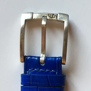 May include: A close-up of a silver watch buckle with the inscription "U.I.N." on the top. The buckle is attached to a blue leather watch strap.