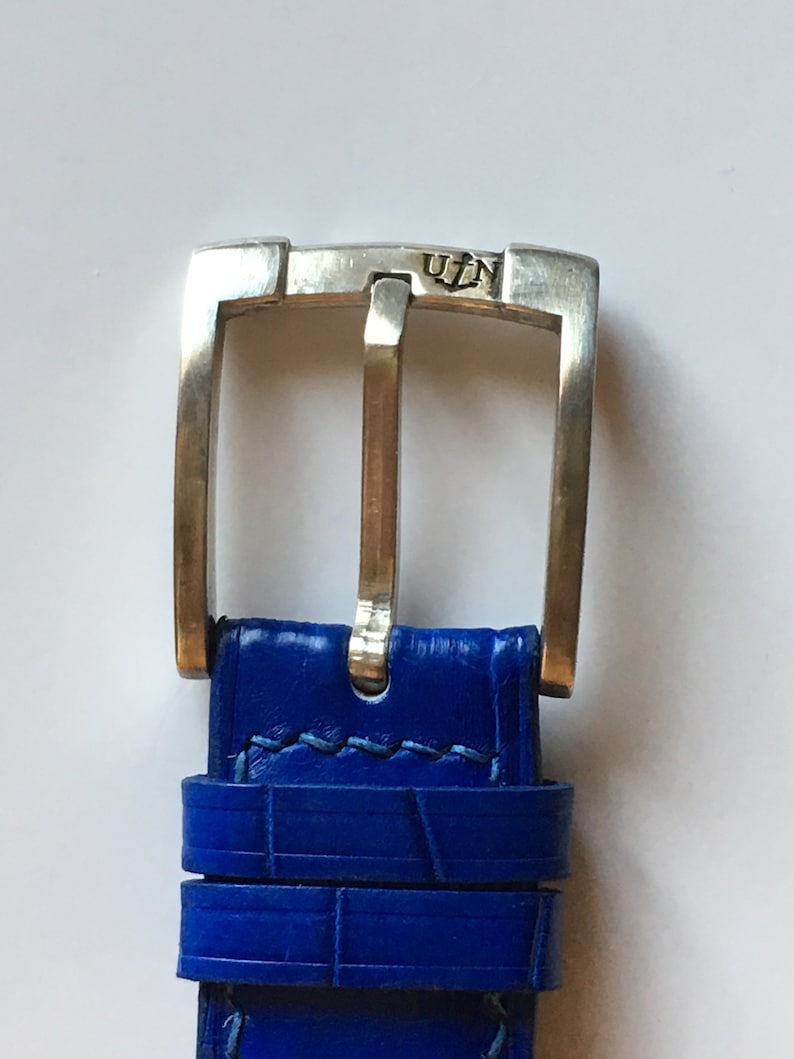 May include: A close-up of a silver watch buckle with the inscription "U.I.N." on the top. The buckle is attached to a blue leather watch strap.