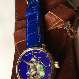 May include: A silver wristwatch with a blue face and a ship illustration. The watch face reads "Ulysse Nardin" and "Santa Maria". The watch has a blue leather strap.