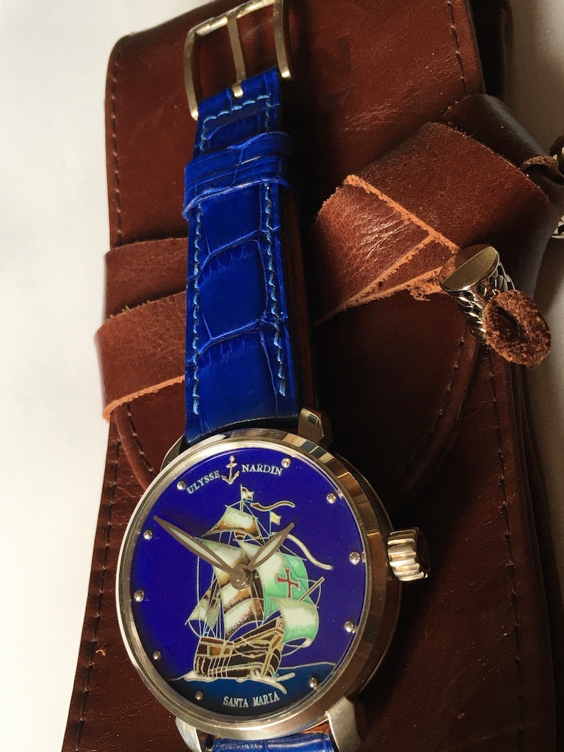 May include: A silver wristwatch with a blue face and a ship illustration. The watch face reads "Ulysse Nardin" and "Santa Maria". The watch has a blue leather strap.
