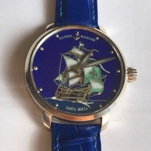 May include: A silver wristwatch with a blue face and a blue leather strap. The face features a painted image of a ship called "Santa Maria" with the text "Ulysse Nardin" above it.