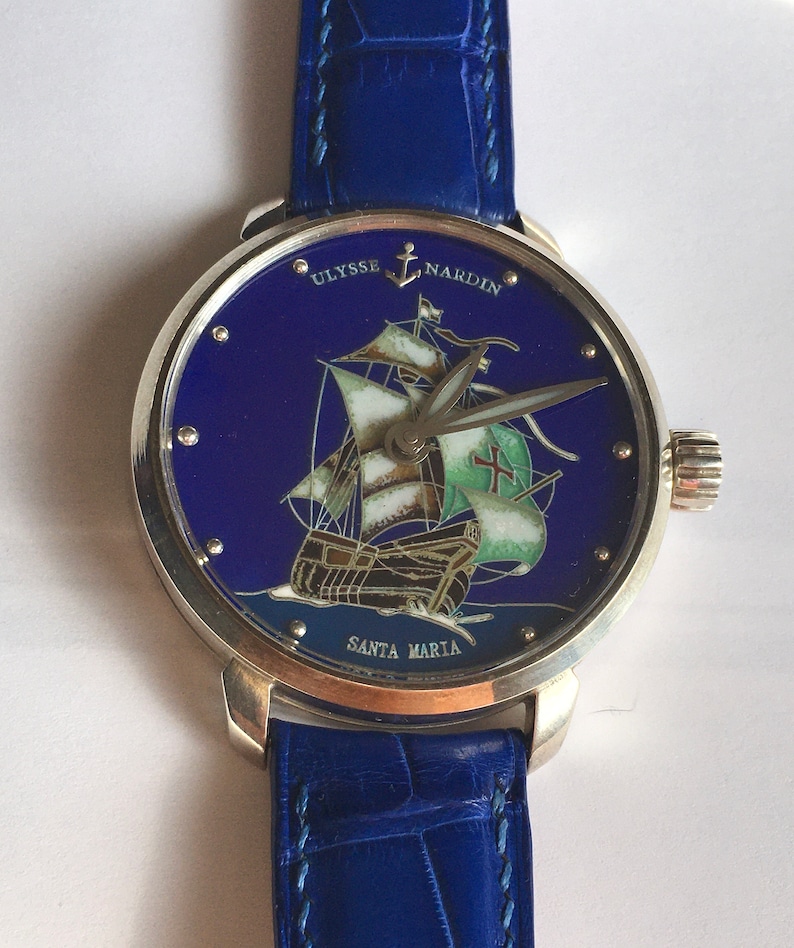 May include: A silver wristwatch with a blue face and a blue leather strap. The face features a painted image of a ship called "Santa Maria" with the text "Ulysse Nardin" above it.
