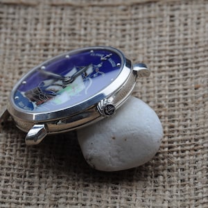 May include: A silver wristwatch with a blue face featuring a ship and the text "Pierre Langier" on the face. The watch is resting on a white rock.