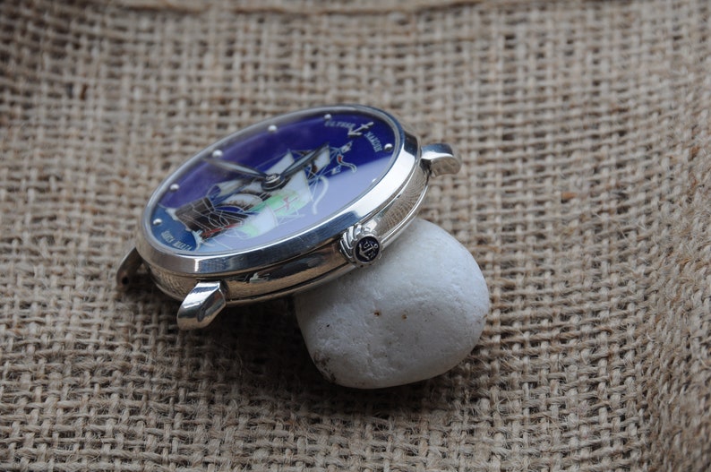 May include: A silver wristwatch with a blue face featuring a ship and the text "Pierre Langier" on the face. The watch is resting on a white rock.