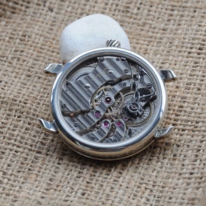 May include: A silver watch with a clear back revealing the intricate inner workings of the movement. The watch is lying on a brown hessian surface.