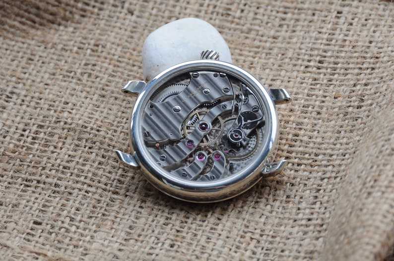 May include: A silver watch with a clear back revealing the intricate inner workings of the movement. The watch is lying on a brown hessian surface.