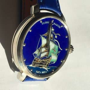 May include: A silver wristwatch with a blue face and a depiction of a ship called "Santa Maria". The watch face also includes the text "Ulysse Nardin". The watch has a blue leather strap.