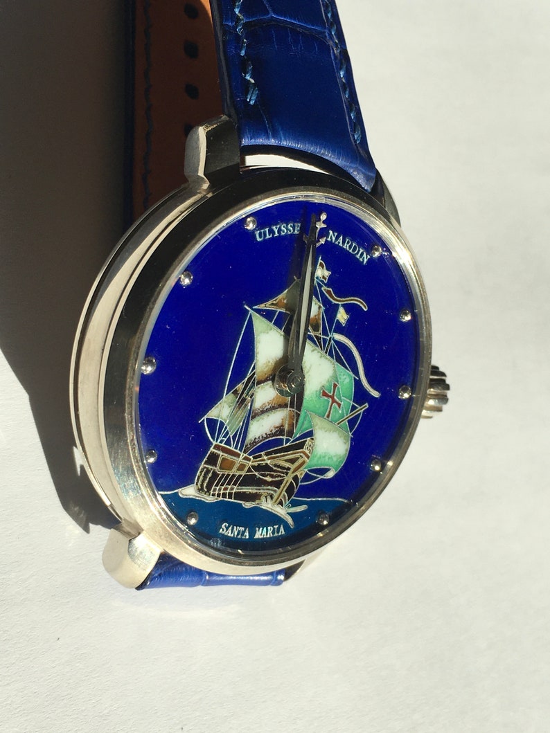 May include: A silver wristwatch with a blue face and a depiction of a ship called "Santa Maria". The watch face also includes the text "Ulysse Nardin". The watch has a blue leather strap.