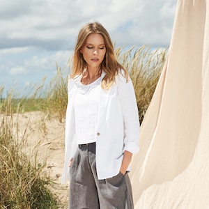 May include: A woman wearing a white linen blazer, a white linen top, gray linen shorts, and gray sandals. She is standing on a sandy beach with a white linen sheet behind her.