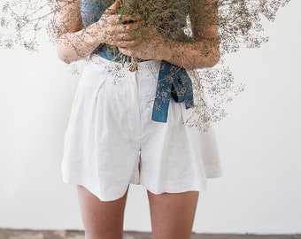 May include: A woman wearing a blue sleeveless top and white linen shorts holds a bouquet of dried flowers. The flowers are arranged in a fan shape and are a light brown color.
