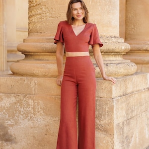 Peut inclure: Une femme portant un haut court et un pantalon large en lin couleur rouille. La tenue est un ensemble deux pièces avec un col en V et des manches courtes évasées. Elle porte des sandales à talons dorées.