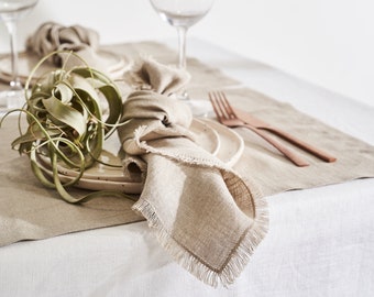 May include: A table setting with a beige linen runner, plates, napkins, and copper-colored forks. Two wine glasses are visible. A decorative green plant adds a natural element to the scene. The napkin has a fringed edge.