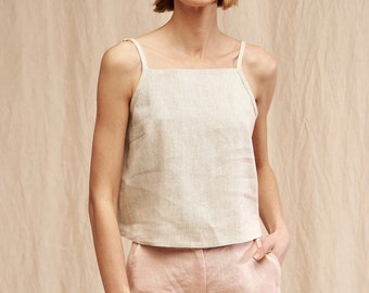 May include: A woman wearing a light beige linen tank top with thin straps and a square neckline. She is also wearing light pink linen pants.