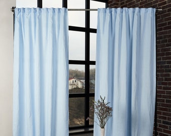 May include: Two light blue linen curtains hanging from a metal rod in front of a window. The curtains are fully extended and the window is behind them. A vase with dried flowers is in front of the curtains.