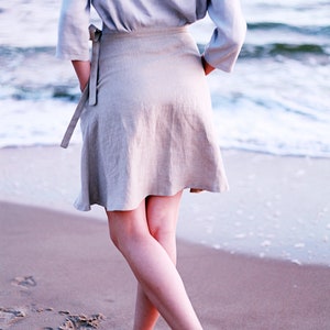 May include: A woman wearing a light blue linen top and a beige linen skirt stands on a sandy beach with the ocean in the background.