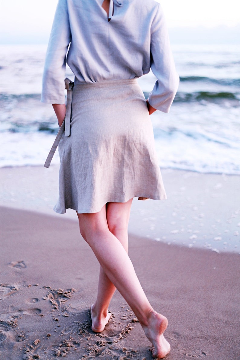 May include: A woman wearing a light blue linen top and a beige linen skirt stands on a sandy beach with the ocean in the background.