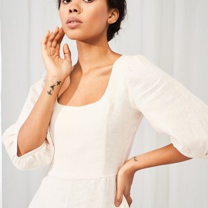 May include: A woman wearing a white linen dress with a square neckline and puffed sleeves. She is standing in front of a white background.