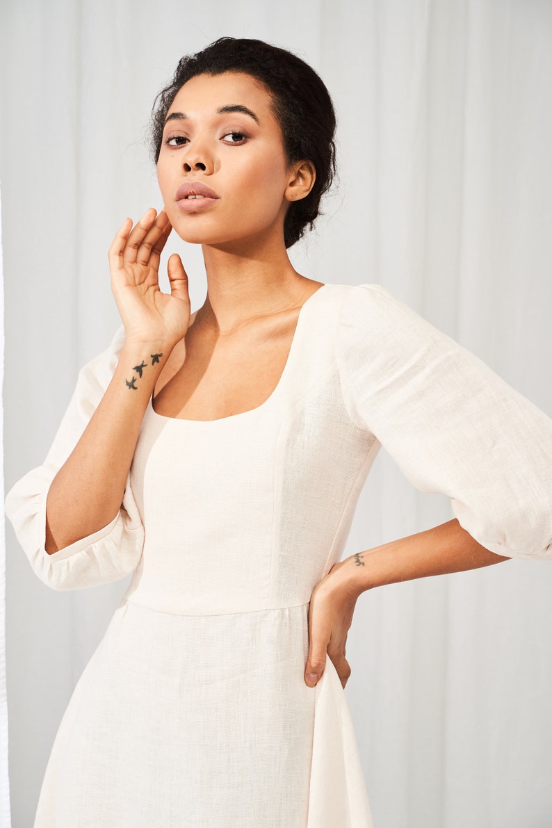 May include: A woman wearing a white linen dress with a square neckline and puffed sleeves. She is standing in front of a white background.