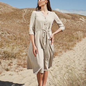 May include: A woman wearing a beige linen dress with a tie belt and three-quarter sleeves stands in a sandy landscape. She is looking to the right of the frame.